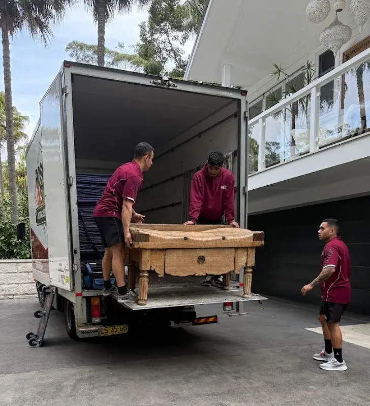 Two Posch & Silva movers lifting a timber table into the back of a truck