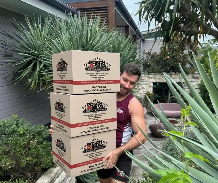 A Posch & Silva mover carrying a stack of branded moving boxes through a garden